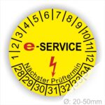 Prüfplaketten rund, Farbe- Gelb, Text "E-SERVICE" in Schwarz danach ein Elektro- Pfeil in Rot, Startjahr- 28, Monatszahlen Oben und Jahreszahlen Unten in Kreisform. Über den Jahreszahlen Text "Nächster Prüftermin"