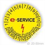 Prüfplaketten rund, Farbe- Gelb, Text "E-SERVICE" in Schwarz danach ein Elektro- Pfeil in Rot, Startjahr- 29, Monatszahlen Oben und Jahreszahlen Unten in Kreisform. Über den Jahreszahlen Text "Nächster Prüftermin"