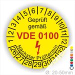 Prüfplaketten rund Farbe- Gelb, Text "Geprüft gemäß VDE 0100" in Schwarz danach ein Elektro- Pfeil in Rot, Startjahr- 26, Monatszahlen Oben und Jahreszahlen Unten in Kreisform. Über den Jahreszahlen Text "Nächster Prüftermin"