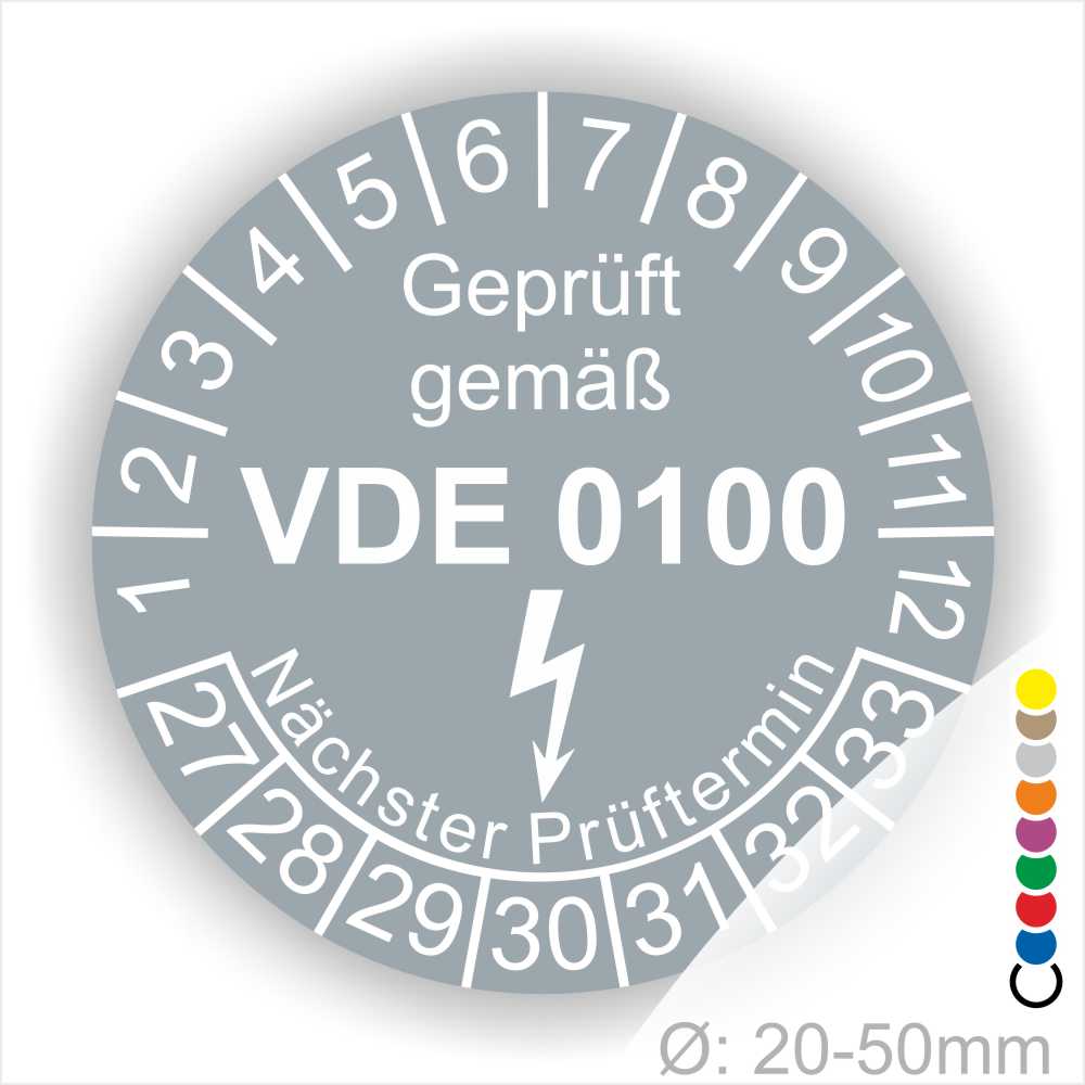 Prüfaukleber rund Farbe- Grau, Text "Geprüft gemäß VDE 0100" in Weiß danach ein Elektro- Pfeil in Weiß, Startjahr- 27, Monatszahlen Oben und Jahreszahlen Unten in Kreisform. Über den Jahreszahlen Text "Nächster Prüftermin"