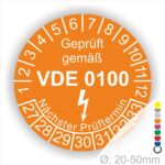 Prüfaukleber rund, Farbe- Orange, Text "Geprüft gemäß VDE 0100" in Weiß danach ein Elektro- Pfeil in Weiß, Startjahr- 27, Monatszahlen Oben und Jahreszahlen Unten in Kreisform. Über den Jahreszahlen Text "Nächster Prüftermin"