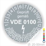 Prüfetikette rund, Farbe- Grau, Text "Geprüft gemäß VDE 0100" in Weiß danach ein Elektro- Pfeil in Weiß, Startjahr- 28, Monatszahlen Oben und Jahreszahlen Unten in Kreisform. Über den Jahreszahlen Text "Nächster Prüftermin"