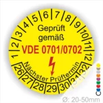 Gelbes Prüflabel mit der Aufschrift „Geprüft gemäß VDE 0701/0702“, das den nächsten Prüftermin anzeigt. Das Label hat einen Durchmesser von 20-50 mm und enthält eine Blitzsymbolik zur Kennzeichnung elektrischer Sicherheit.