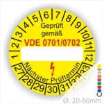Gelbes Prüfplakett mit dem Text "Geprüft gemäß VDE 0701/0702" und Platz für das nächste Prüftermin-Datum. Das Plakett hat einen Durchmesser von 20-50 mm und zeigt eine Liste von Prüfterminen von 1 bis 33. Ideal für die Kennzeichnung elektrischer Geräte.